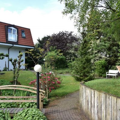 Ein Haus mit rotem Dach und Gaube im Hintergrund. Im Vordergrund ist ein gepflegter Garten mit einer Holzbrücke über einem kleinen Graben. Rechts auf der Wiese stehen ein Strandkorb und eine weiße Bank unter Bäumen.
