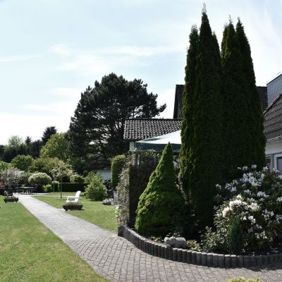 Ein Garten mit einem gepflasterten Weg, gesäumt von Büschen und hohen Koniferen. Rechts ist ein modernes Haus mit dunklem Dach, links eine Wiese mit Sitzbank. Bäume und blauer Himmel im Hintergrund.