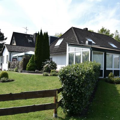 Großes Einfamilienhaus mit schwarzen Dachschindeln und Wintergarten auf der rechten Seite. Es steht auf einem gepflegten, leicht ansteigenden Rasen mit verschiedenen Sträuchern und Bäumen, umgeben von einem Holzzaun im Vordergrund.