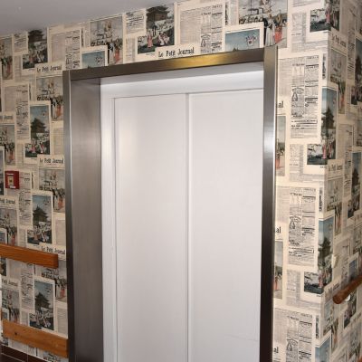 Ein geschlossener Aufzug mit weißen Türen ist von einer Wand umgeben, die mit Tapete im Design alter Zeitungsseiten bedeckt ist. Rechts und links der Tür verlaufen braune Handläufe. Der Boden besteht aus braunen Fliesen.