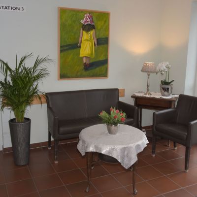 Ein Wartebereich mit zwei braunen Sesseln und einem Sofa um einen runden Tisch mit weißer Tischdecke. Auf dem Tisch steht ein Blumenarrangement. An der Wand hängt ein Bild eines Kindes in gelber Kleidung. Pflanzen schmücken den Raum.