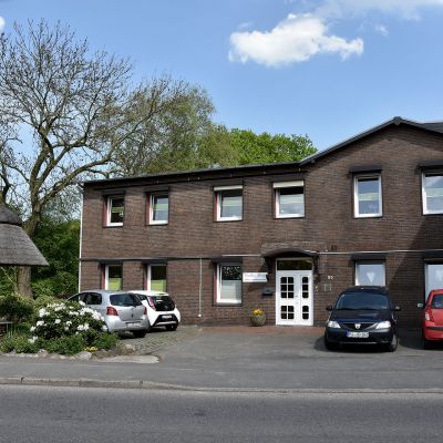Ein zweistöckiges Backsteingebäude mit weißen Fensterrahmen und einer weißen Eingangstür. Vor dem Gebäude sind drei geparkte Autos und ein Brunnen mit Strohdach. Links steht ein Baum mit grünem Laub. Der Himmel ist blau mit einigen Wolken.