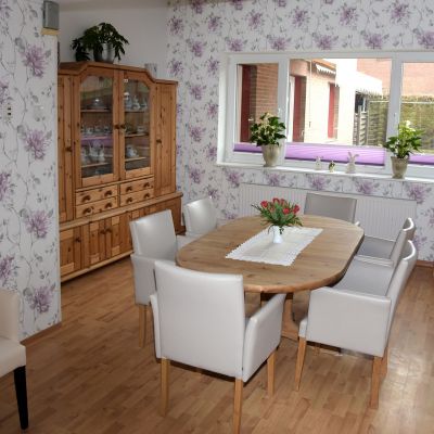 Esszimmer mit Holzfußboden, ovalem Holztisch und sechs weißen Stühlen. Links ein Holzschrank mit Glastüren und Spiegel darüber. Rechts ein Fenster mit Pflanzen und eine Kommode, darüber hängt ein Fernseher. Wände mit floralem Muster.