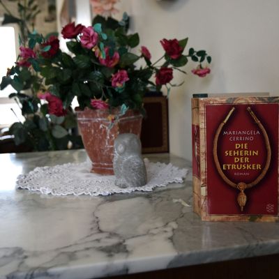 Ein Buch mit rotem Einband steht auf einem Marmortisch. Daneben befinden sich ein Topf mit Blumen und eine kleine, verzierte Figur auf einem Häkeldeckchen. Im Hintergrund ist ein Fenster und ein Sessel zu sehen.