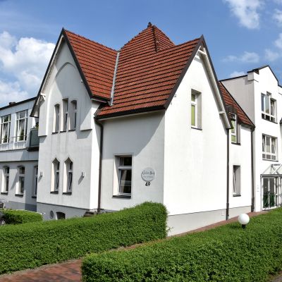 Ein weißes Gebäude mit roten Dächern und mehreren Dachgauben. Vor dem Gebäude sind eine Hecke, ein Gehweg und einige parkende Autos. Der Himmel ist blau mit einigen Wolken.