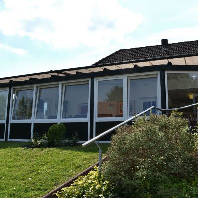 Ein modernes Gebäude mit großen Fensterfronten, die von einem dunklen Rahmen umgeben sind. Darunter ist eine grüne Rasenfläche mit Büschen. Der Himmel ist teilweise bewölkt und blau.