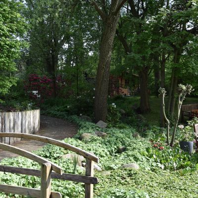 Ein Garten mit üppigem Grün, Bäumen und Blumen. Ein geschwungener Holzsteg führt über einen Weg. Links steht eine Lampe mit weißem globusförmigem Schirm, rechts sind Bänke auf einer Rasenfläche. Im Hintergrund sind dichte Bäume.